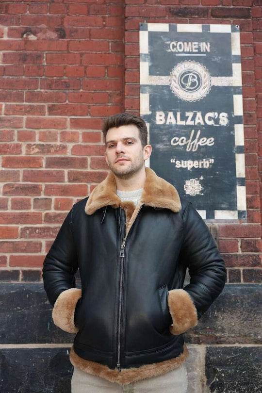 Man wearing Classic Ginger Brown B3 Bomber Aviator Shearling Jacket with fur trim standing in front of a brick wall with a Balzac's Coffee sign.