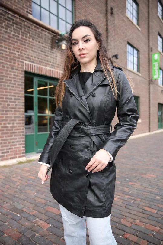 Woman wearing a black trench coat standing in front of a brick building.