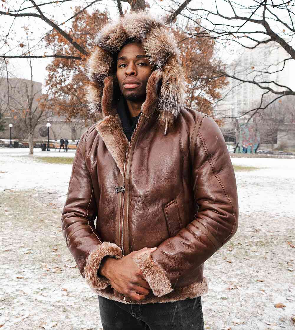 Man wearing a brown hooded bomber leather jacket with fur trim in a snowy park setting.