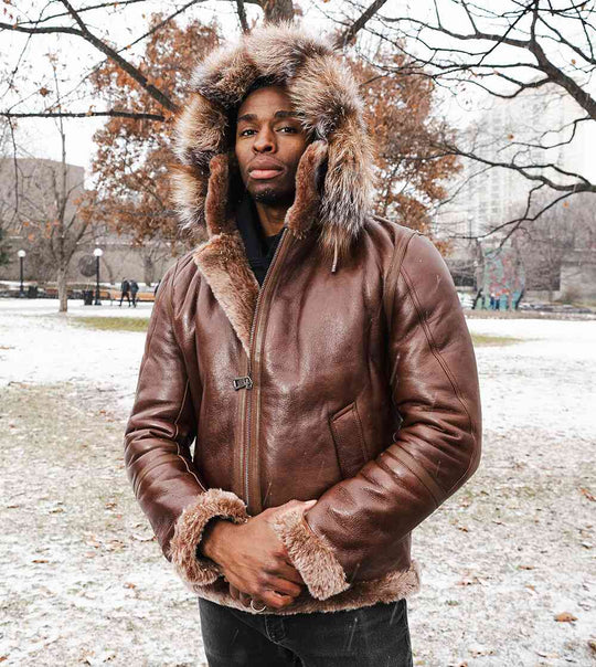 Man wearing a brown hooded bomber leather jacket with fur trim in a snowy park setting.