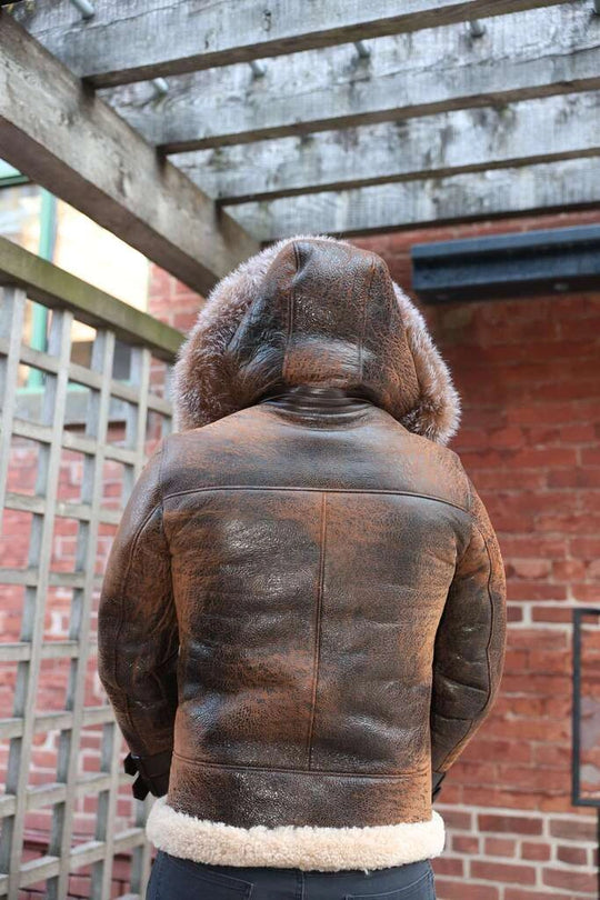 Back view of Rocco vintage brown aviator shearling jacket with fur hood worn by a person, standing in front of a brick wall.