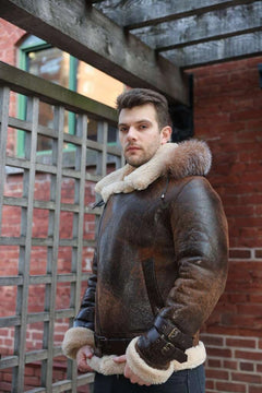 Man wearing Rocco vintage brown aviator shearling jacket with fur trim zips closed standing in front of a brick wall.
